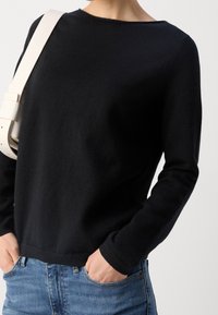 Czarny dzianinowy sweter z okrągłym dekoltem, długimi rękawami i luźnym krojem. Noszony z niebieskimi dżinsami, ręce w kieszeniach, oraz jasnokolorową torebką na ramię.