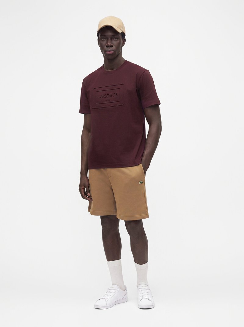 Мъж, носещ бежова шапка, бордова тениска Lacoste, светлокафяви къси панталони, бели чорапи и бели спортни обувки, стоящ на бял фон.
