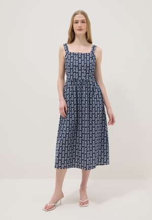 Frau steht und trägt ein marineblaues ärmelloses Midikleid mit weißen geometrischen Mustern und weißen Sandalen mit Absatz vor schlichtem Hintergrund.