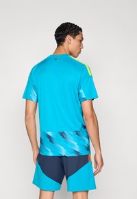 Man facing away wearing a bright blue sports shirt with geometric patterns and matching blue and navy shorts.