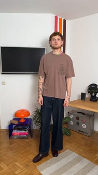 T-shirt gráfica castanho, calças de ganga azuis largas e sapatos bordô. Em pé sobre um chão de madeira com uma lâmpada colorida e uma planta nas proximidades.