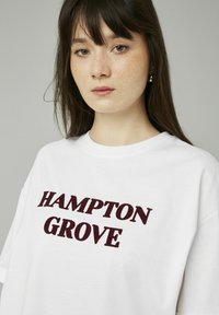 T-shirt blanc avec inscription en relief maroon "HAMPTON GROVE" sur le devant. Coupe classique avec manches courtes et col rond.