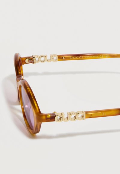 Lunettes de soleil rondes translucides marron avec logo Gucci doré sur les branches et verres teintés, posées sur une surface blanche.