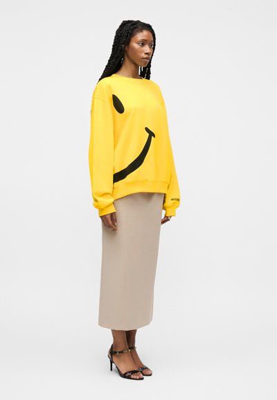 Femme aux longs cheveux tressés, portant un sweat-shirt jaune avec un smiley noir, une jupe midi beige et des sandales à talons noires, debout de profil.
