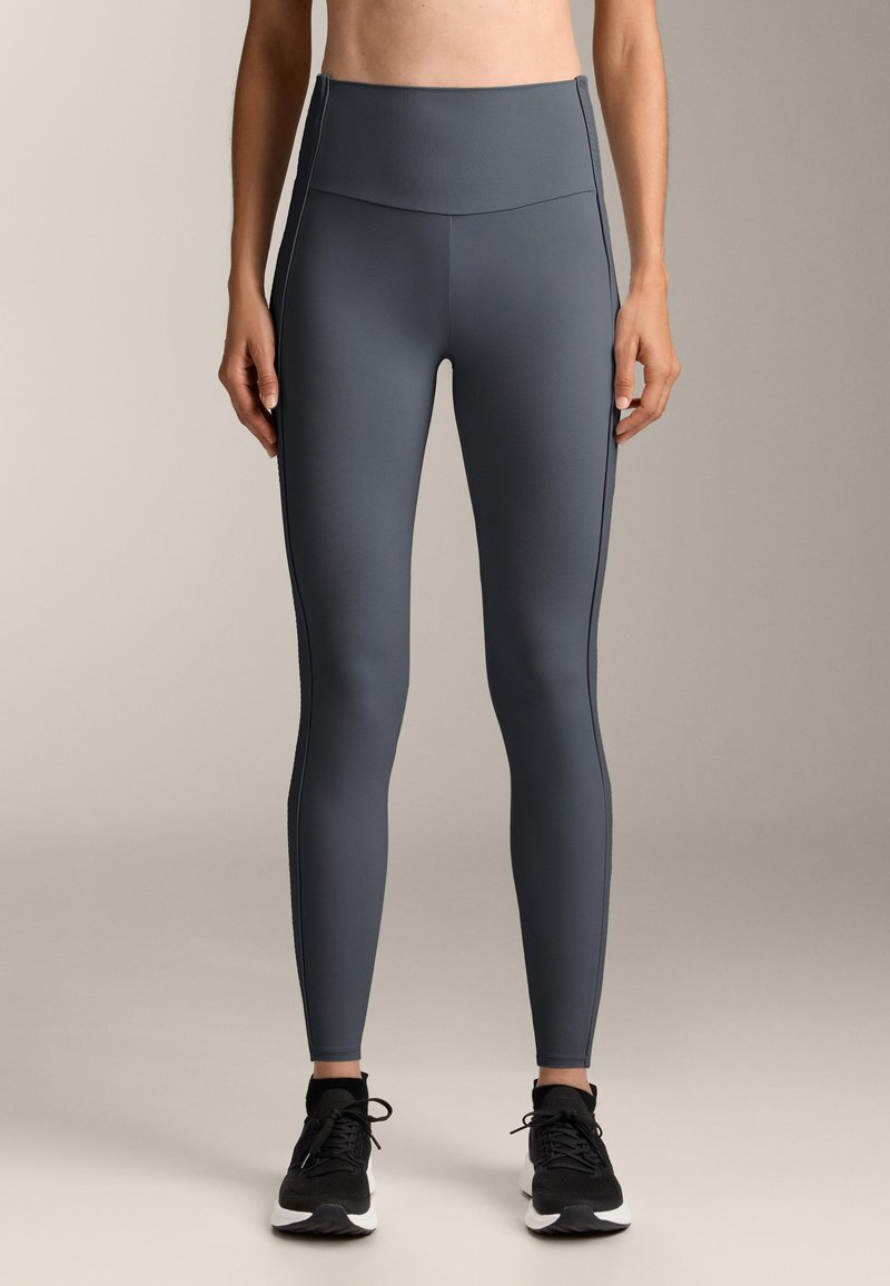 Leggings griși cu talie înaltă, confecționate dintr-un material neted și elastic, cu cusături contrastante, design mulat și croială până la glezne. Se poartă încălțăminte sport negre.