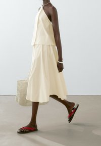 Robe deux pièces côtelée jaune clair avec un col halter ; associée à des sandales marron avec des brides rouges. Sac tressé à la main.