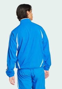 Blue athletic jacket with white accents, full zip, high collar, and elastic cuffs. Smooth texture, lightweight design, suitable for active wear.