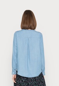 Chemise chambray bleu clair à manches longues et ourlet arrondi. Caractérisée par un pli au dos et des poignets à boutons. Associée à une jupe noire à fleurs.
