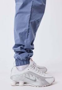 Persoon in blauwe joggingbroek en witte Nike sneakers met zichtbare luchtkussens op een effen lichte achtergrond.