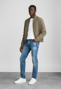 Olivinėje žalioje medžiagoje bomber tipo striukė, derinama su balta marškinėliais, šviesiai mėlynomis džinsomis ir baltais sportbačiais; paprastas dizainas, prigludusi forma.