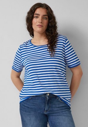 Kobieta o długich kręconych włosach, ubrana w niebiesko-białą koszulkę w paski z krótkim rękawem oraz niebieskie dżinsy, stojąca przy jednolitej szarej ścianie.