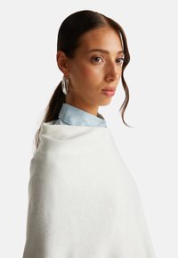 Bílý pletený šál položený na světle modré košilové tričko. Má hladkou strukturu a volný, elegantní design. Viditelné jsou jasné kruhové náušnice.