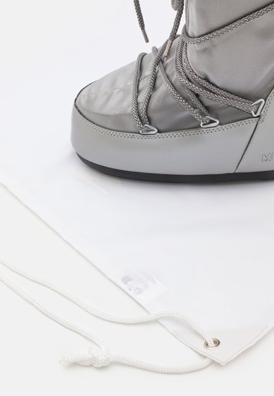 Botte grise imperméable avec surface texturée, embout renforcé et lacets gris. Présentée sur un sac de protection blanc avec fermeture à cordon.