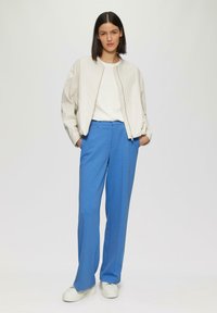 Giacca bomber beige chiaro, maglietta bianca a girocollo, pantaloni larghi blu e sneakers bianche. Tessuto morbido con una texture liscia e un design minimale.