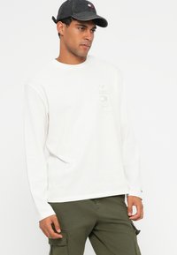 Λευκό, μακρυμάνικο βαμβακερό πουκάμισο με γραφικό τύπωμα στο αριστερό στήθος, συνδυασμένο με ελαιόχρωμα cargo παντελόνια και σκούρο καπέλο.