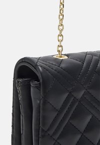 Svart quiltad läderhandväska med en guldkedjestraps och en cirkulär detaljer i metall. Har en texturerad yta och en strukturerad form.
