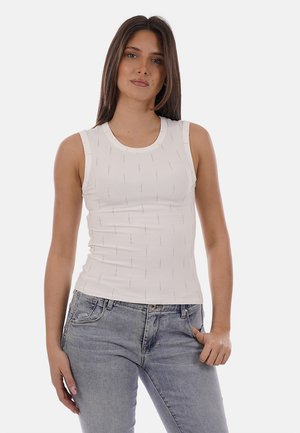 Donna con capelli castani lunghi e lisci, che indossa un top bianco senza maniche aderente e jeans azzurri, in piedi con una mano in tasca.