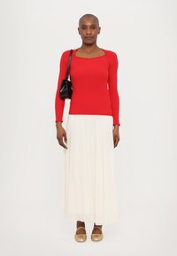 Haut à manches longues rouge côtelé, jupe maxi plissée blanche et ballerines en jute dorées. Le modèle tient un petit sac à main noir. Lignes simples et épurées.