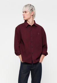 Bordeaux skjorte med knapper og krave, sorte knapper og oprullede ærmer. Har et diskret logo på brystet, parret med mørke denimjeans.