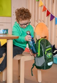 Grüner Kinder-Hoodie, schwarze Jeans, hält eine blau gemusterte Wasserflasche, benutzt einen grün-grauen Rucksack mit Dinosaurier-Design.