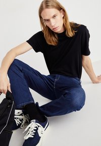 Camiseta de algodón negra, jeans de mezclilla azul con costuras visibles y zapatillas de deporte marinas con detalles blancos. Estilo informal, corte relajado.