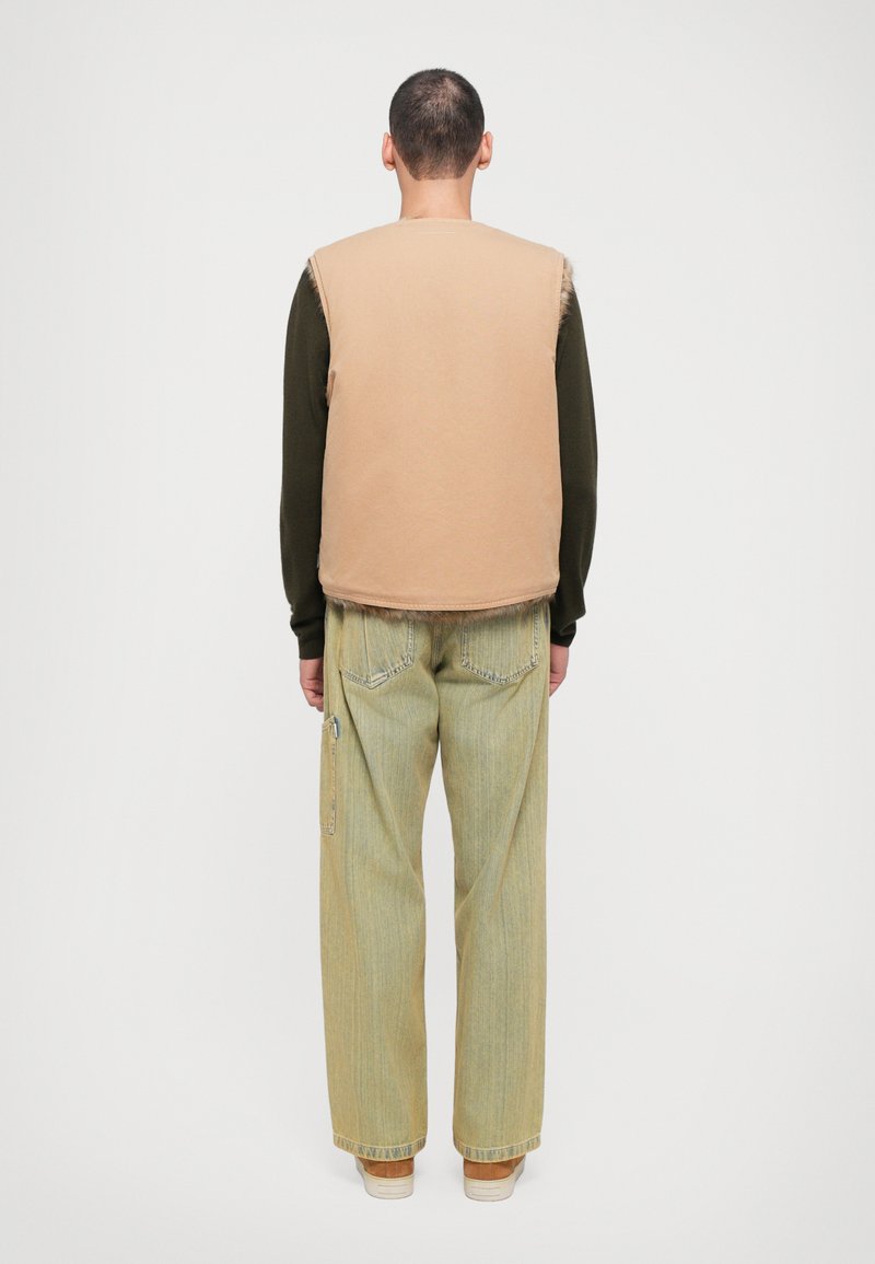 Beige sleeveless vest over a dark long-sleeve shirt, paired with light green wide-leg jeans. Features raw edges and side pocket zipper.