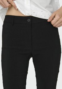 Pantalon noir avec une texture lisse, doté d'une fermeture à bouton, d'une coupe ajustée et d'une taille haute. La ceinture comporte des passants de ceinture.