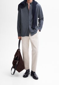 Camicia a manica lunga grigio scuro, pantaloni color crema, stivali in pelle nera, e una borsa in suede marrone con tracolla regolabile, maglione lavorato a maglia blu navy appoggiato.