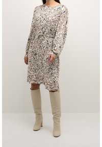 Robe beige avec un motif abstrait gris et noir, manches longues, taille cintrée, accompagnée de bottes beige montant jusqu'aux genoux.