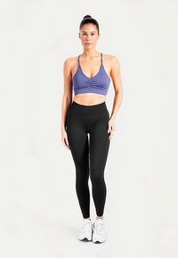 Soutien-gorge de sport violet avec détails froncés, associé à un legging noir taille haute. Le mannequin porte des chaussures de sport blanches, debout devant un fond neutre.