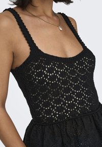 Top in maglia nera con una texture a motivo, caratterizzato da spalline sottili e una sezione inferiore svasata. Una delicata collana d'oro accentua il design.