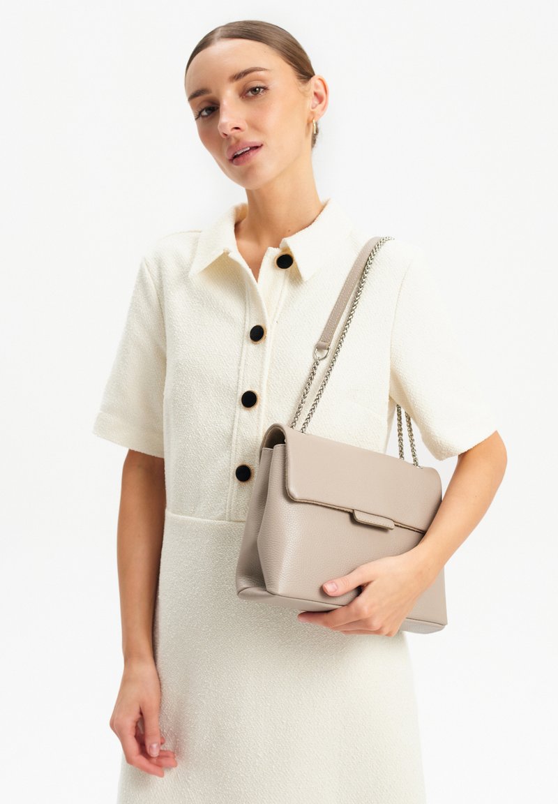 Borsa a mano in pelle beige chiaro con un design strutturato, chiusura a pattina e tracolla a catena. Presentata con un outfit bianco testurizzato e bottoni neri.
