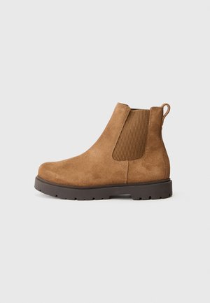 Bottines Chelsea en daim beige avec panneau latéral élastique et languette sur une semelle crantée marron foncé, présentées de profil sur fond blanc.