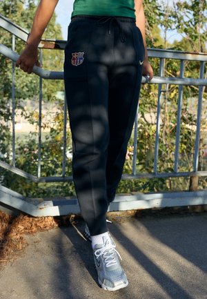 Persoon draagt zwarte FC Barcelona trainingsbroek en grijze Nike sneakers, staand buiten bij een metalen reling.