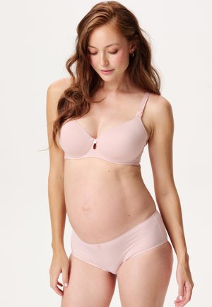 Mujer embarazada con cabello largo, vistiendo un sujetador premamá rosa claro y ropa interior a juego, mirando hacia abajo con una expresión suave.