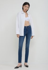 Jeune femme portant un top blanc court, une chemise oversized blanche, un jean skinny taille haute bleu et des escarpins à talons kitten noirs, se tenant contre un mur blanc.