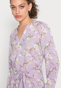 Vestido floral de lavanda com mangas compridas, decote em V e faixa na cintura. Apresenta flores brancas e folhas verdes em um tecido texturizado.