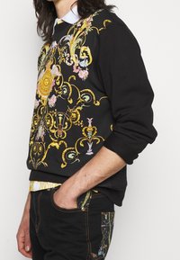 Black sweatshirt featuring vibrant floral and ornate gold patterns, with ribbed cuffs and neckline, worn over patterned pants.
