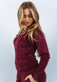 Bordowa, dopasowana bluza na zamek wykonana z miękkiego materiału, z boczną kieszenią zapinaną na guzik oraz ściągaczami na rękawach, zestawiona z pasującymi spodnimi.