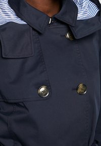 Granatowy płaszcz podwójnie zapinany na duże guziki w kolorze musztardy, z niebiesko-białym paskowanym wykończeniem kołnierza, noszony przez osobę.