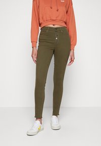 Jean skinny vert olive avec taille haute, fermeture à cinq boutons, et ourlets effilochés aux chevilles, associés à un sweat à capuche court corail et des baskets blanches.