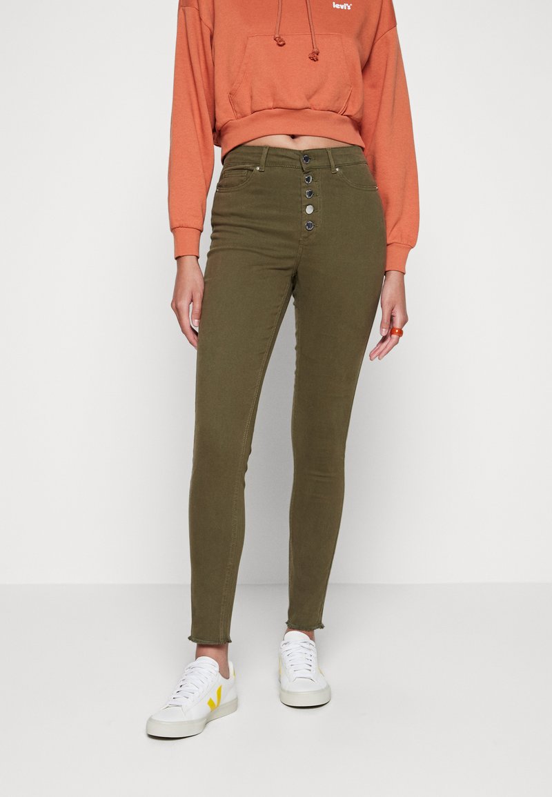 Jean skinny vert olive avec taille haute, fermeture à cinq boutons, et ourlets effilochés aux chevilles, associés à un sweat à capuche court corail et des baskets blanches.