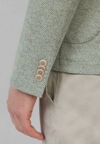 Jasnozielona marynarka z tkaniny tweed w kolorowe wzory. Posiada cztery jasne guziki oraz dopasowany krój, pokazana w połączeniu z spodniami w neutralnym odcieniu.
