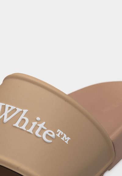 Παντόφλα σε μπεζ χρώμα με λευκό ανάγλυφο κείμενο "White™" στο λουρί, με απλό και ομαλό σχεδιασμό.