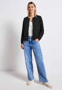 Černá prošívaná bomber bunda s kulatým výstřihem, v kombinaci se světle modrými širokými džíny a bílými teniskami. Jednoduchý design, bez viditelných vzorů.