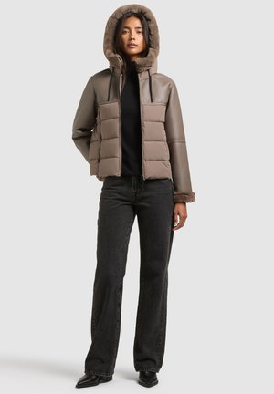 Femme portant une veste matelassée marron avec capuche et poignets doublés de fourrure, un top noir, un jean noir et des bottes noires pointues, debout sur un fond uni.
