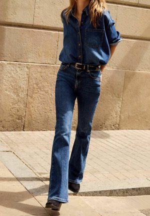 Chemise en denim bleu foncé avec fermeture à boutons et poche poitrine, associée à un jean évasé bleu foncé et des bottines noires.