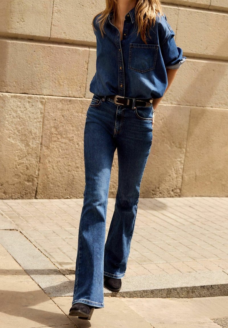 Camisa de mezclilla en azul oscuro con cierre de botones y bolsillo en el pecho, combinada con pantalones vaqueros acampanados en azul oscuro y botas negras hasta el tobillo.