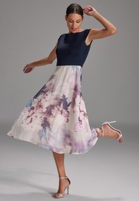 Robe ajustée bleu marine avec un corsage et une jupe plissée crème ornée de grands motifs floraux roses et violets. Associée à des talons argentés à lanières.