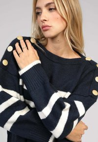 Pull bleu marine avec des rayures blanches, doté de grands boutons dorés sur l'épaule et de poignets côtelés, fabriqué en maille épaisse.